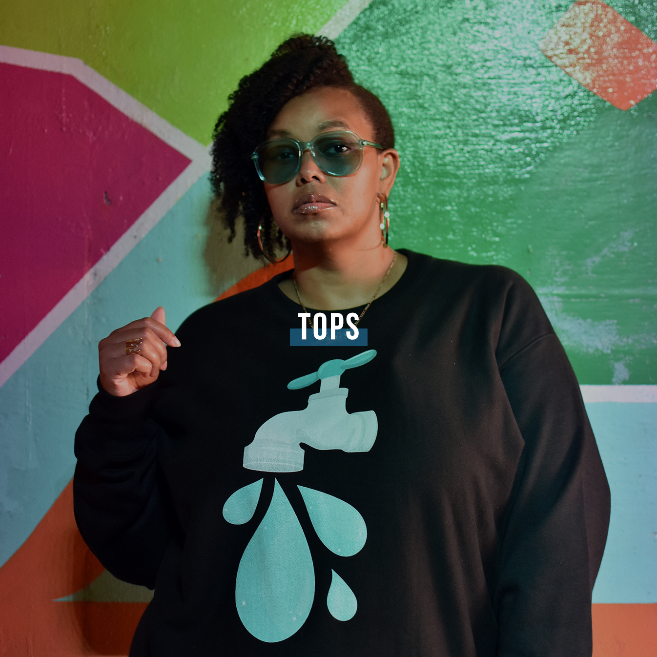 Waist up shot of a Black woman wearing the 1550 Brand Big Drip Champion sweatshirt, which features an illustrated image of a faucet with large drops of water falling from it. Her hair is curly and piled on one side and she is wearing turquoise sunglasses to match the image on the sweatshirt.